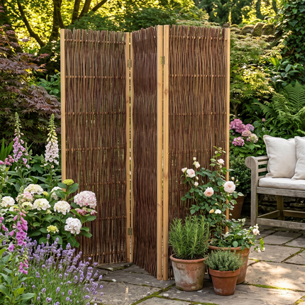 A three-panel bamboo folding screen stands on a stone patio in a lush garden setting, with potted plants, a wooden bench, and greenery in the background.