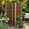 A three-panel bamboo folding screen stands on a stone patio in a lush garden setting, with potted plants, a wooden bench, and greenery in the background.