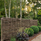 A natural wattle privacy fence made of interlaced branches, planted in a landscaped garden bed with green shrubs and plants, next to a gravel path.