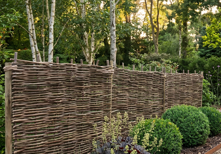 A natural wattle privacy fence made of interlaced branches, planted in a landscaped garden bed with green shrubs and plants, next to a gravel path.