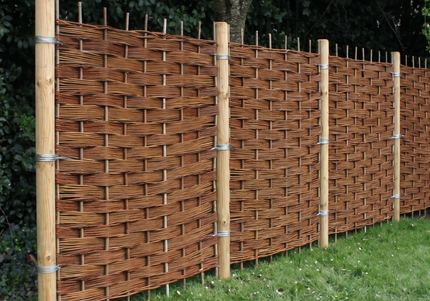 A natural wicker fence panel with vertical wooden posts and metal bands, shown installed outdoors on a grassy lawn with trees in the background.