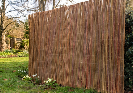 A tall, rectangular bamboo or reed privacy screen panel standing on a green lawn in a backyard setting, with trees and shrubs in the background.